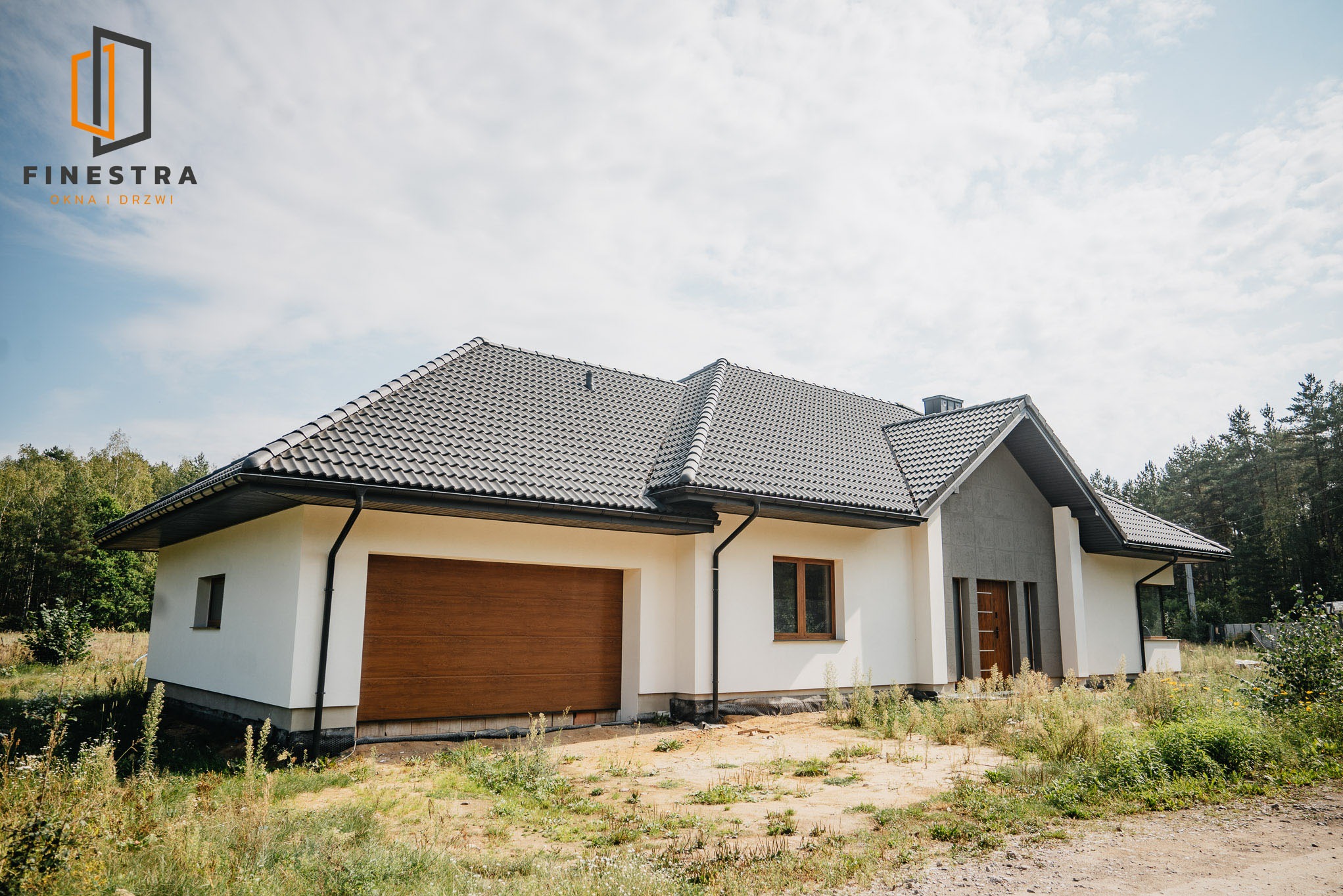Nowoczesny dom jednorodzinny z brązową bramą garażową segmentową i oknami w kolorze drewna, na tle zieleni i błękitnego nieba. Widoczny fragment elewacji i dachu.