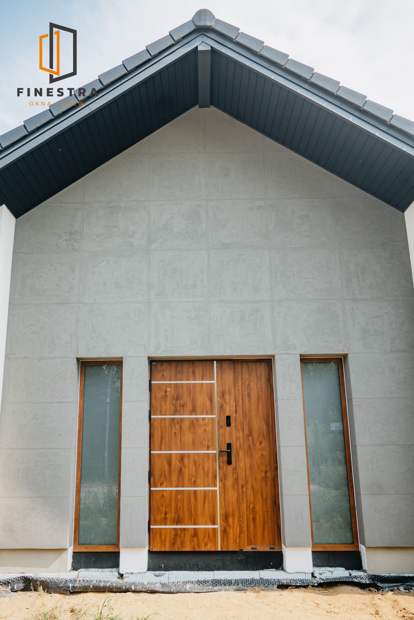 Nowoczesny dom z drewnianymi drzwiami wejściowymi i wąskimi oknami po bokach. Elewacja w odcieniach szarości, dach dwuspadowy z ciemnej dachówki.