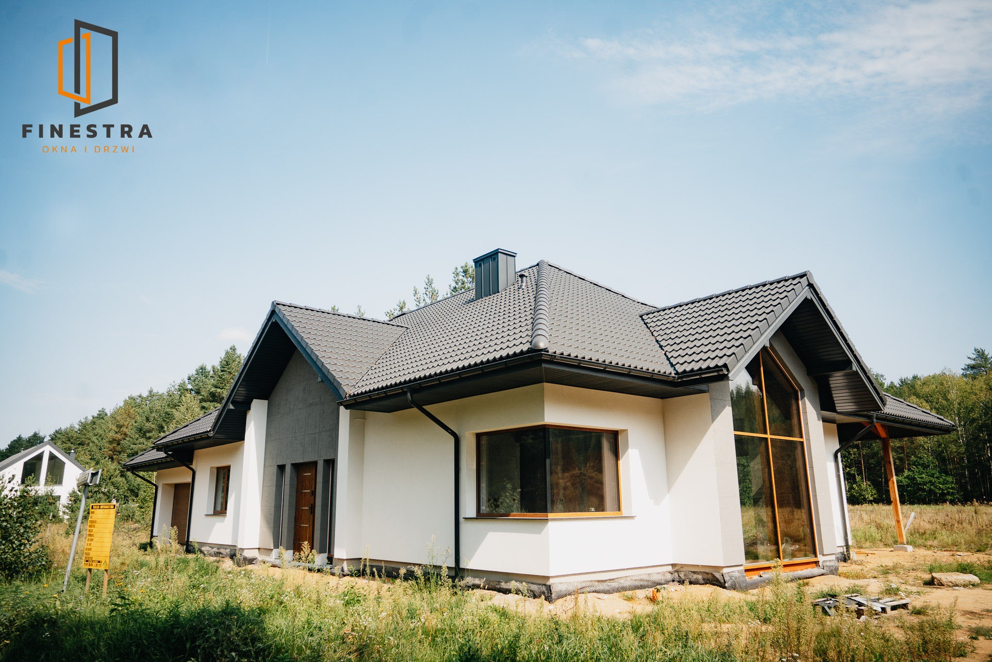 Nowoczesny dom jednorodzinny z dużymi oknami i szarą dachówką, otoczony zielenią. Widoczny fragment elewacji z oknami i drzwiami wejściowymi.