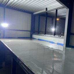 Kms - Świeżo wylana, mokra, szara powierzchnia podłogi w hali przemysłowej, z widocznymi refleksami światła i taśmą zabezpieczającą przy ścianach.