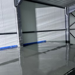 Kms - Świeżo wylana, gładka, szara podłoga w hali przemysłowej, zabezpieczona niebieską folią przy ścianie, z widoczną metalową konstrukcją dachu i ścian.