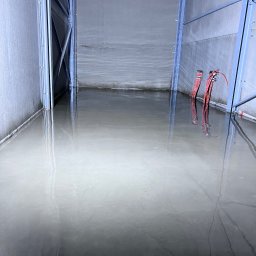 Kms - Świeżo wylana, mokra, szara podłoga w hali przemysłowej, z widocznymi przewodami elektrycznymi na ścianie. Metalowa konstrukcja ramy hali.