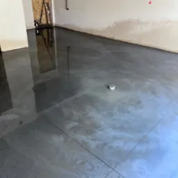 Wypoziomowana, szara posadzka przemysłowa z widocznym odpływem podłogowym i odbiciem narzędzi w tle.