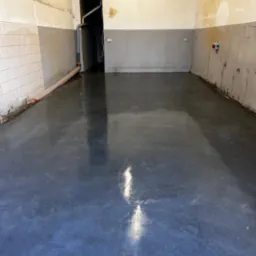 Świeżo wylana, błyszcząca, szara podłoga w pomieszczeniu z surowymi ścianami, widoczne instalacje rurowe.