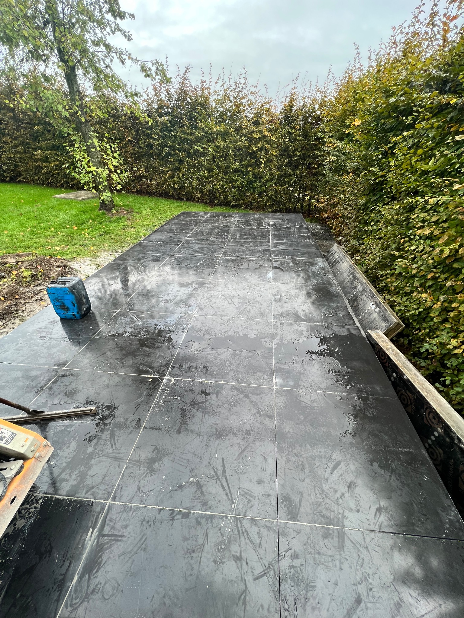 Układanie mokrych, ciemnych płytek na tarasie betonowym w ogrodzie, widoczny niebieski pojemnik i narzędzia do cięcia płytek.