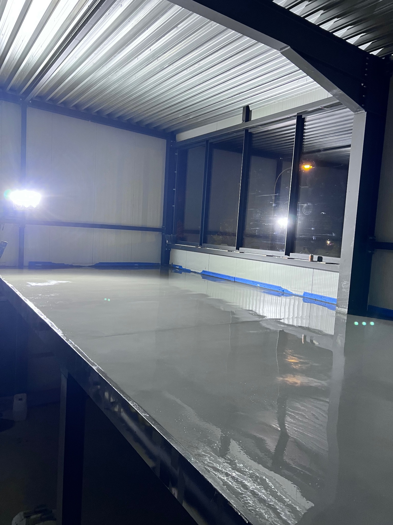 Świeżo wylana, mokra, szara powierzchnia podłogi w hali przemysłowej, z widocznymi refleksami światła i taśmą zabezpieczającą przy ścianach.