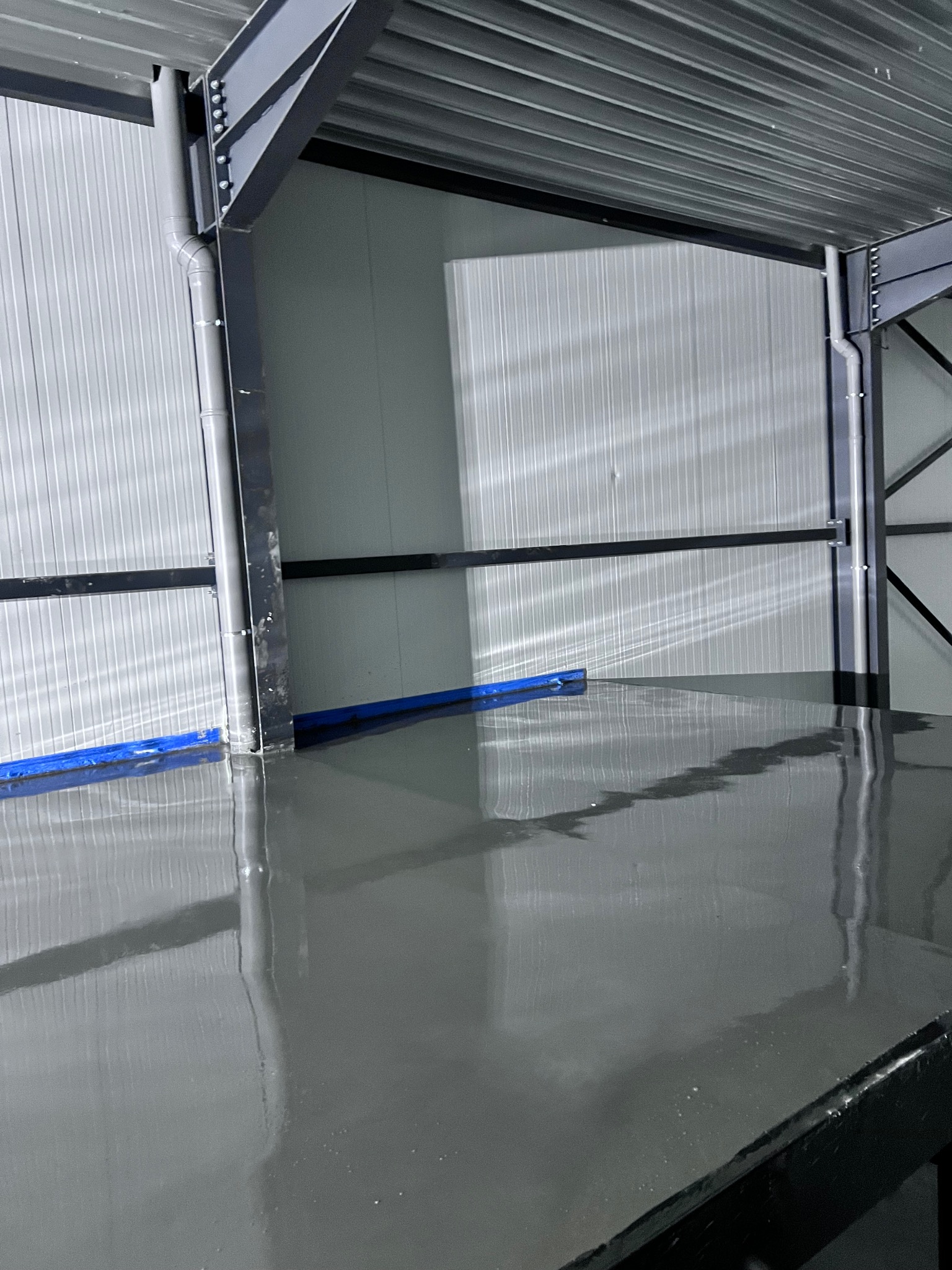 Świeżo wylana, gładka, szara podłoga w hali przemysłowej, zabezpieczona niebieską folią przy ścianie, z widoczną metalową konstrukcją dachu i ścian.