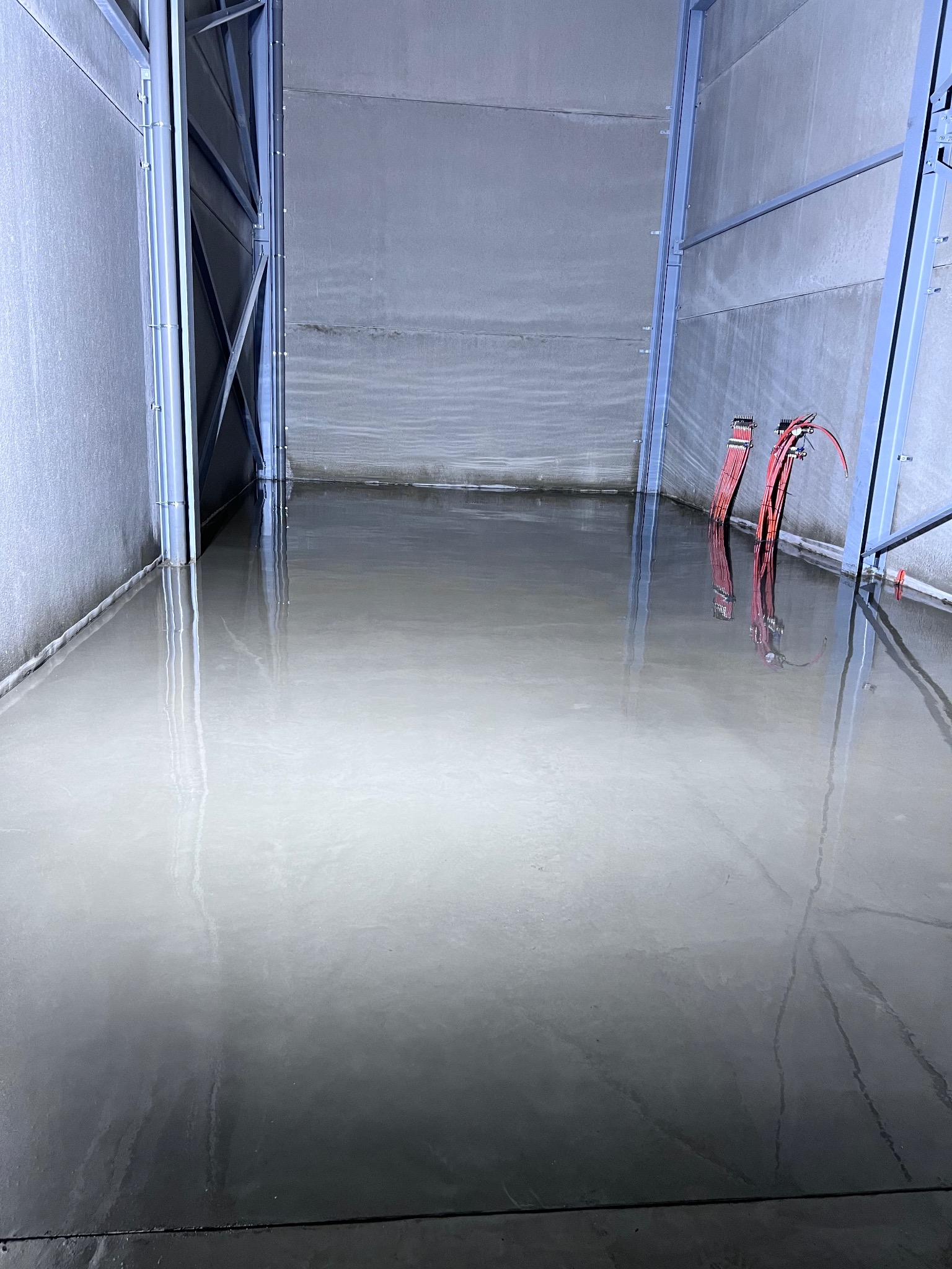 Świeżo wylana, mokra, szara podłoga w hali przemysłowej, z widocznymi przewodami elektrycznymi na ścianie. Metalowa konstrukcja ramy hali.