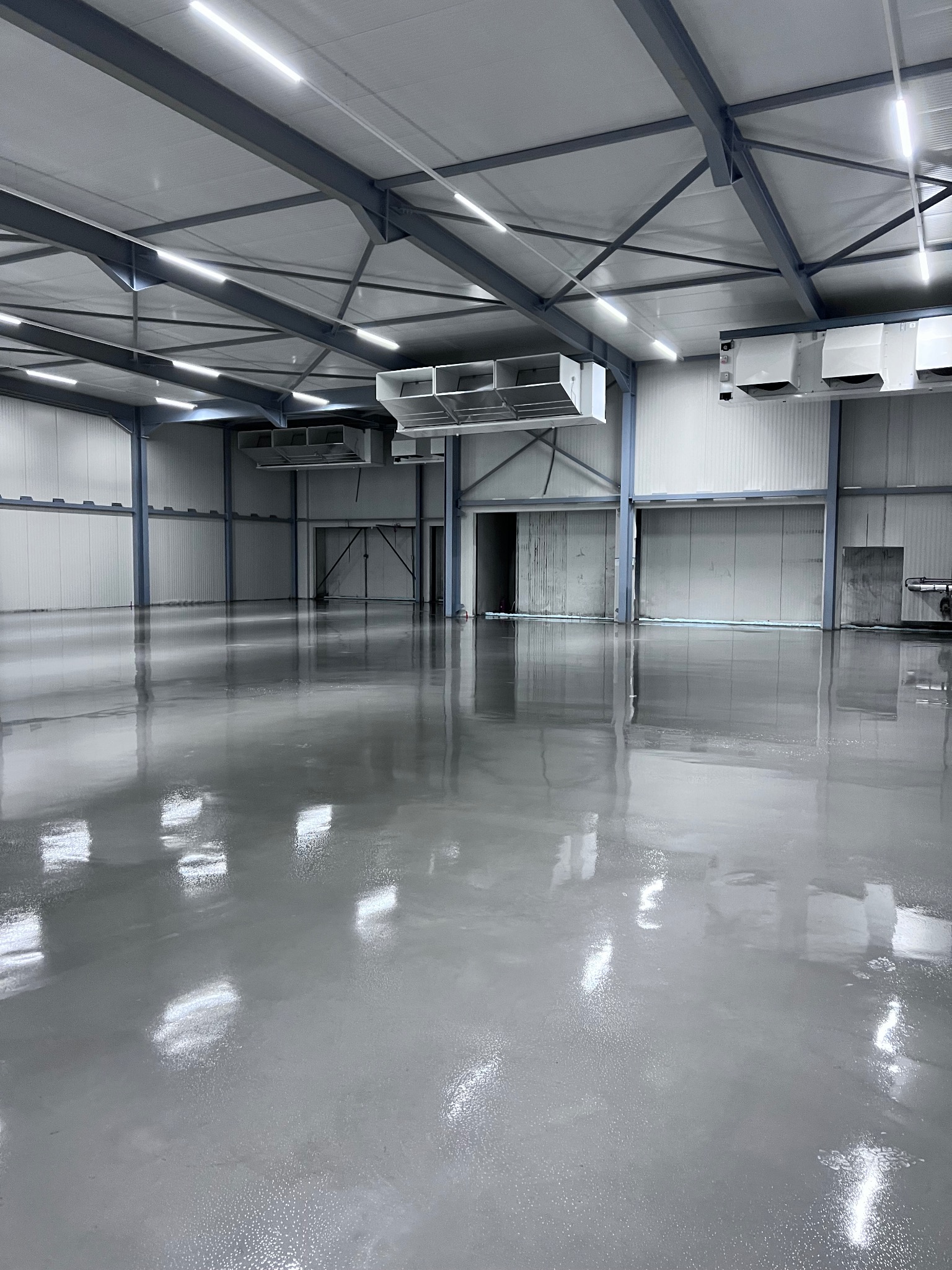 Wnętrze hali przemysłowej z gładką, szarą podłogą, odbijającą światło od lamp sufitowych. Widoczne metalowe konstrukcje stropu i system wentylacji pod sufitem.