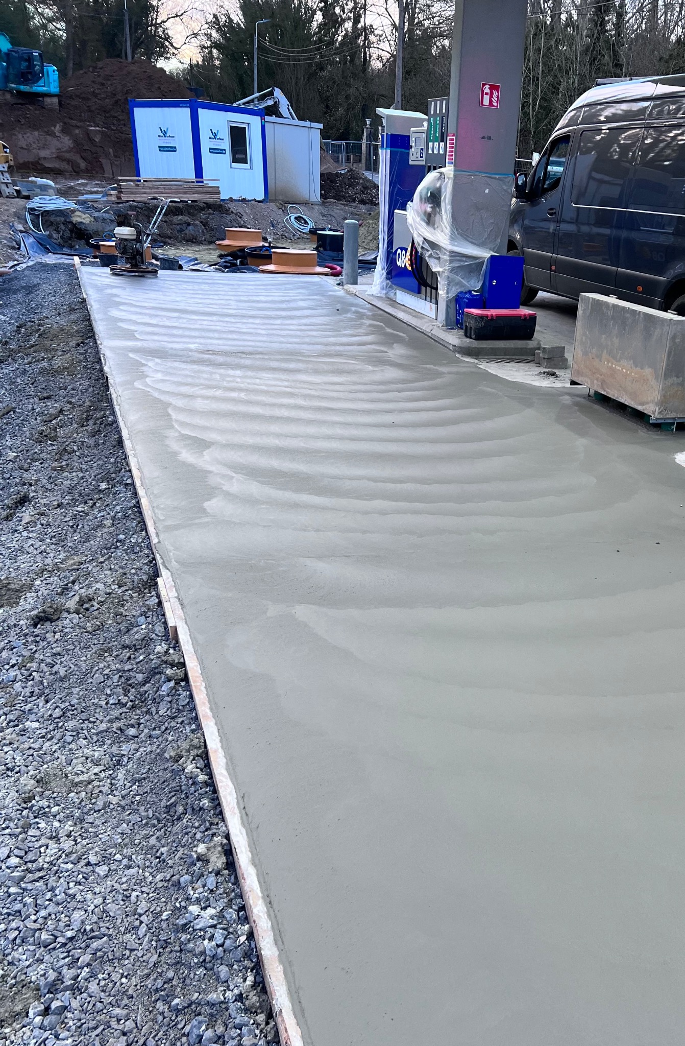 Świeżo wylana, szara powierzchnia przy stacji benzynowej, z widocznymi śladami po zacieraczce mechaniczej, obok zaparkowany van i tymczasowe zaplecze budowy.
