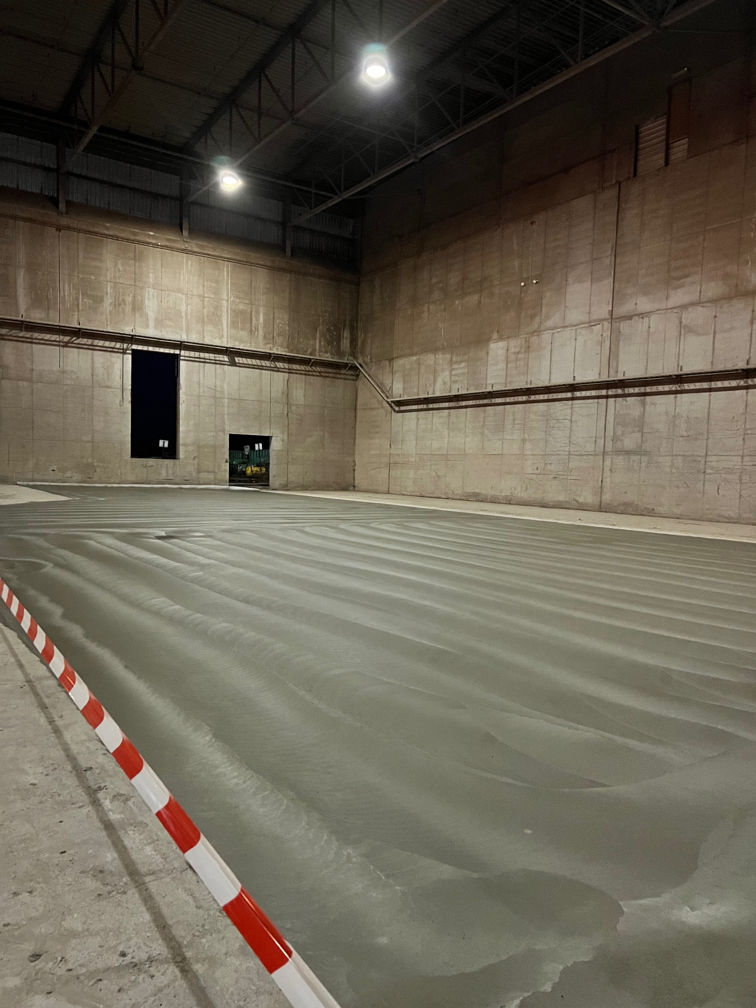 Świeżo wylana, mokra, szara podłoga w hali przemysłowej z widocznymi śladami po narzędziach do rozprowadzania betonu, zabezpieczona biało-czerwoną taśmą ostrzegawczą.