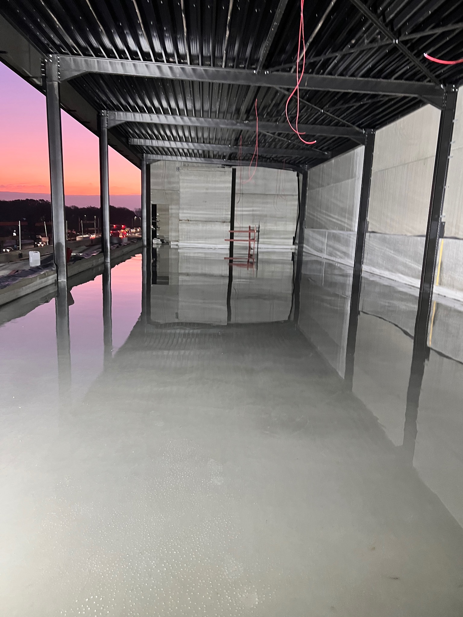 Świeżo wylana, gładka, jasnoszara powierzchnia podłogi w surowym, industrialnym wnętrzu z metalową konstrukcją stropu i słupami; widoczne odbicie zachodzącego słońca w mokrej powierzchni.