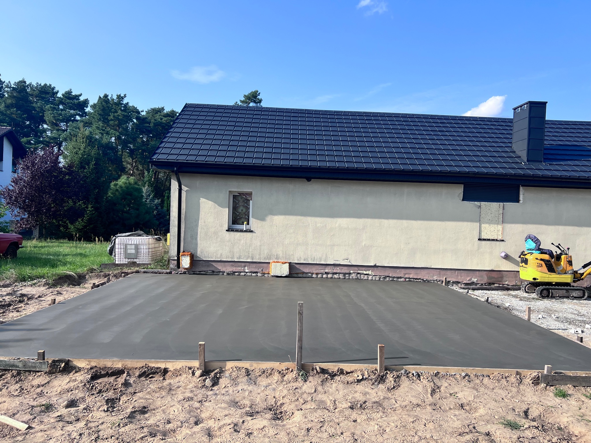 Świeżo wylana, gładka powierzchnia betonowa, przygotowana pod taras, z widocznym szalunkiem drewnianym i minikoparką JCB w tle, na tle domu z ciemną dachówką i błękitnego nieba.