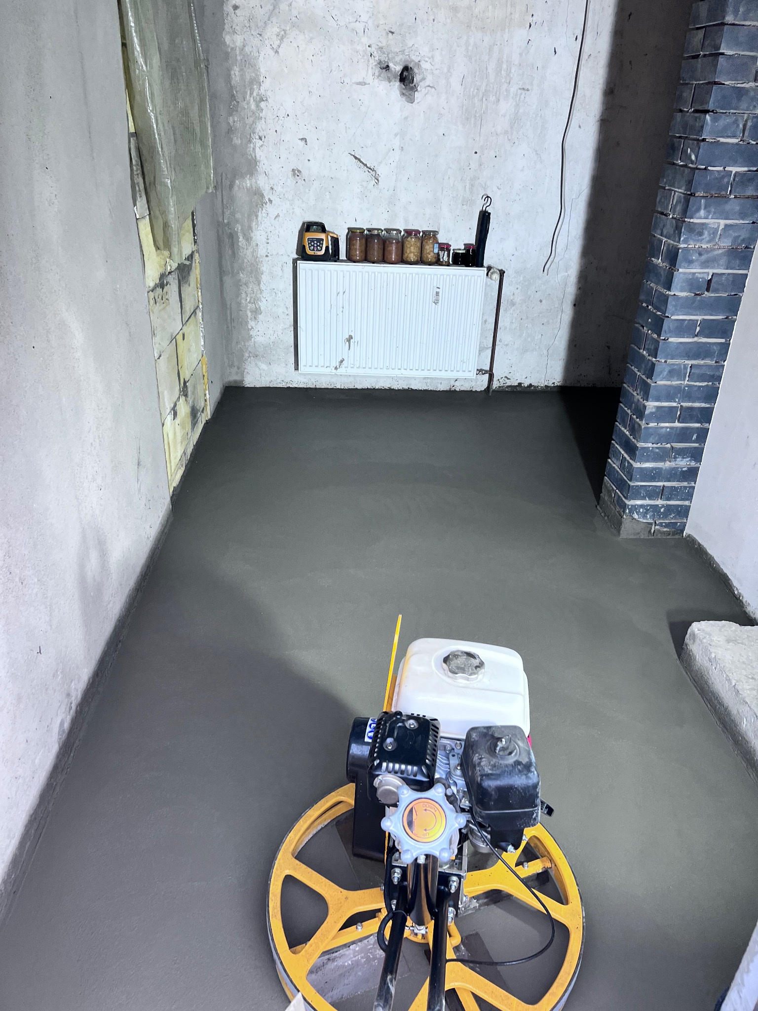 Praca w toku w Gorzowie Wielkopolskim: zacieranie świeżo wylanej, szarej podłogi za pomocą zacieraczki mechanicznej; widoczny fragment ściany z cegły i grzejnik z słoikami na górze.