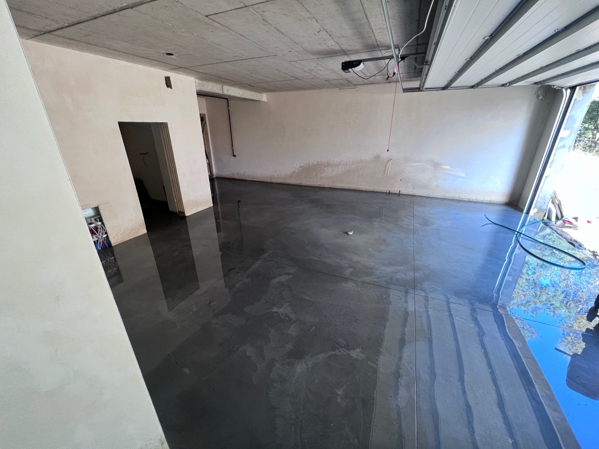 Świeżo wylana, gładka, szara podłoga w surowym pomieszczeniu z betonowym sufitem i otwartą bramą garażową, widoczne ślady wilgoci.