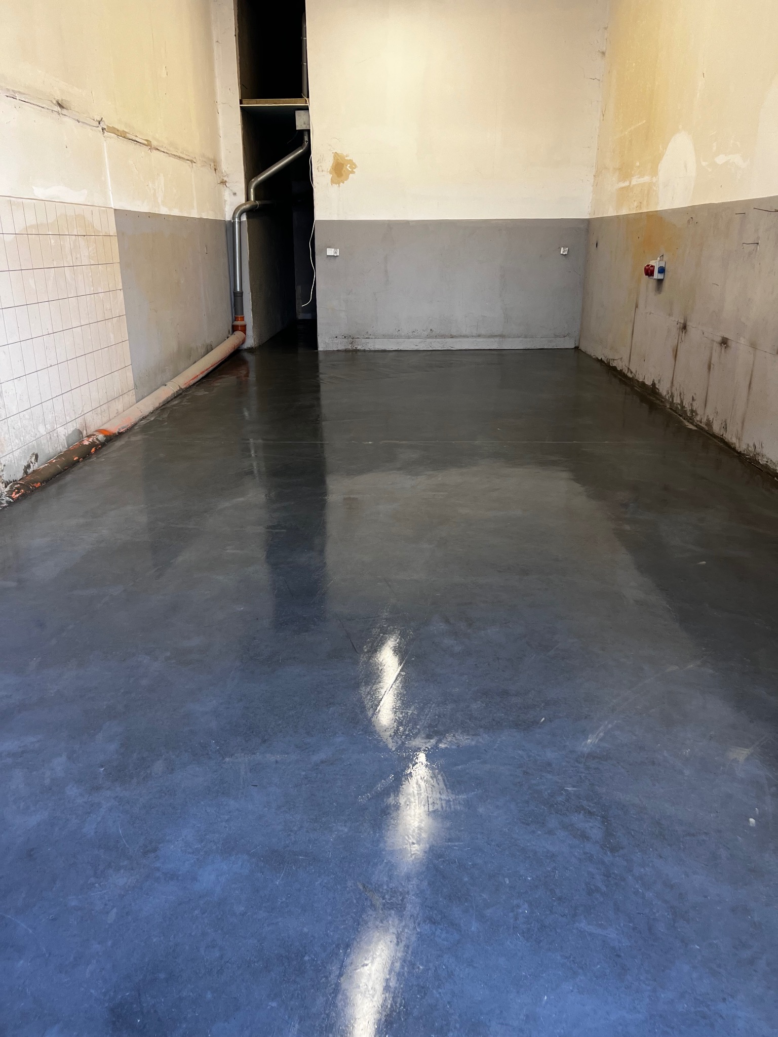 Świeżo wylana, błyszcząca, szara podłoga w pomieszczeniu z surowymi ścianami, widoczne instalacje rurowe.