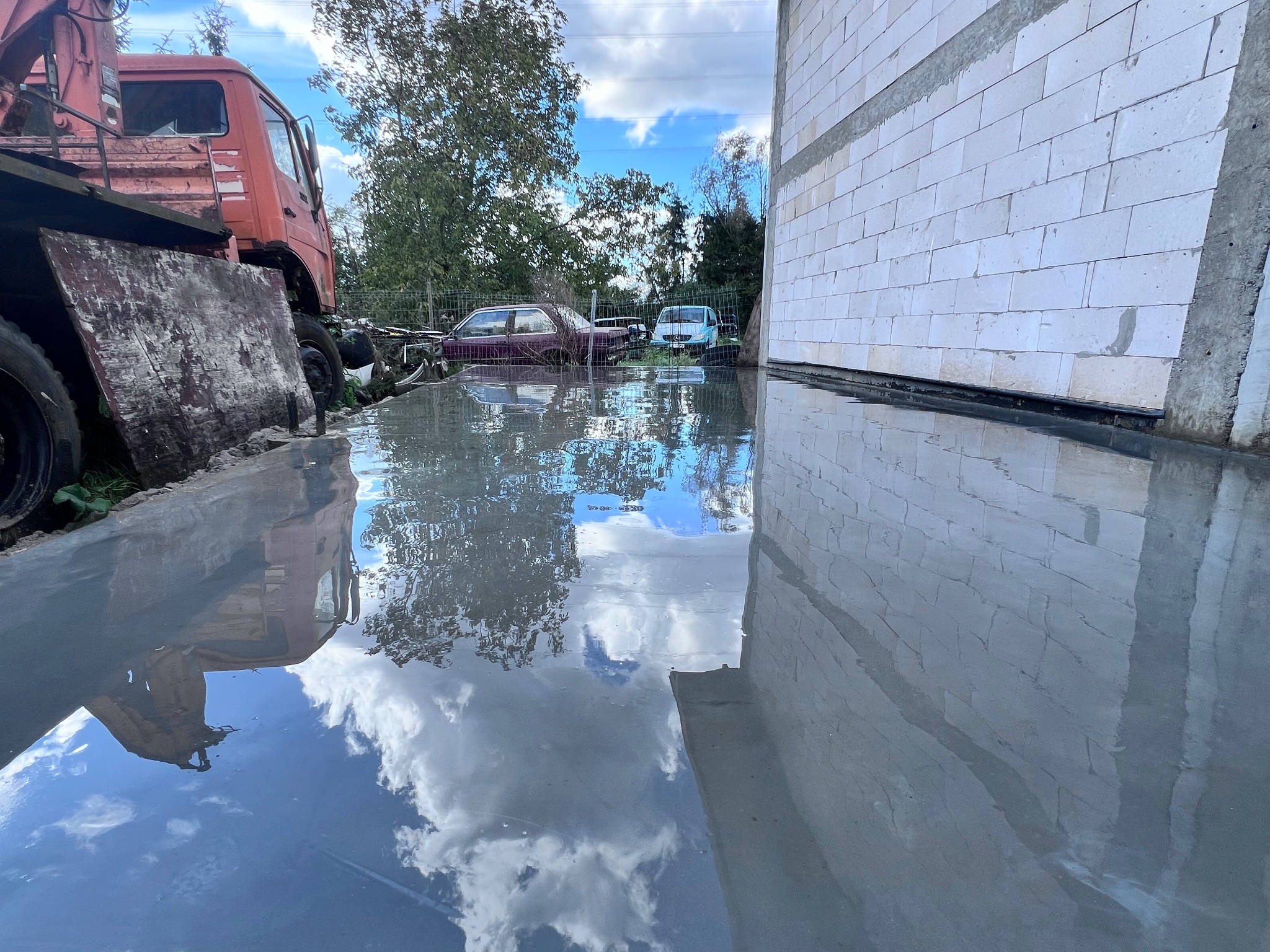 Świeżo wylana, mokra powierzchnia pod budynkiem z białych bloczków, odbijająca niebo i chmury, z widocznym starym dźwigiem w tle.