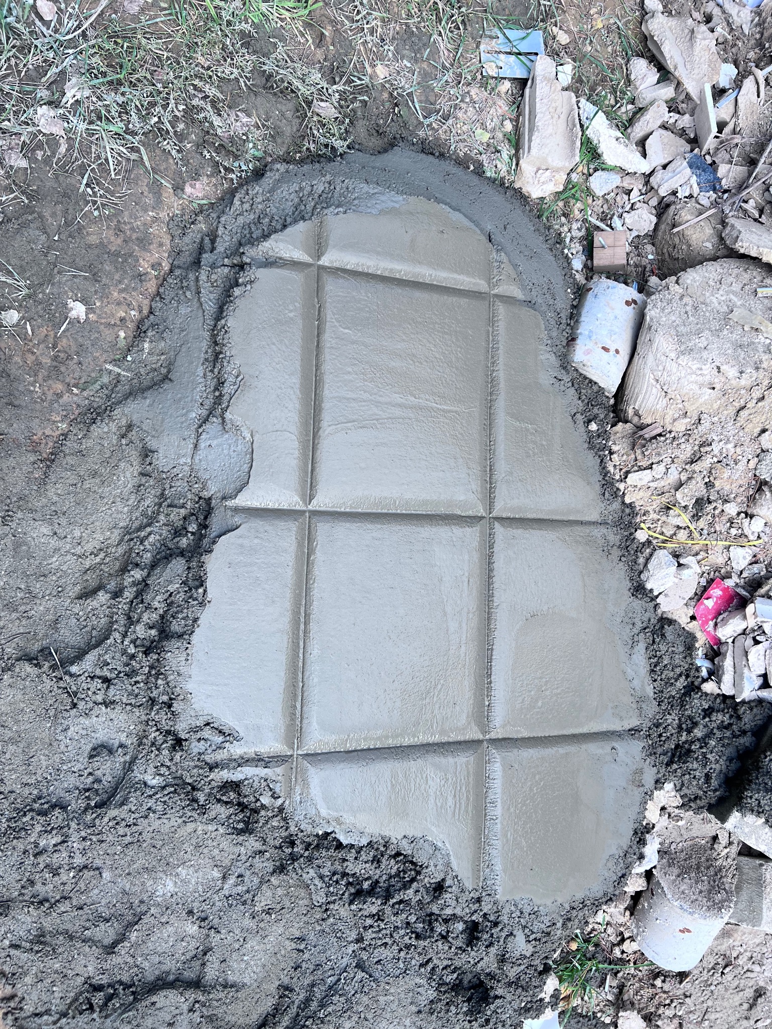 Świeżo wylana, kwadratowa płyta betonowa z nacięciami tworzącymi wzór kratki, otoczona mokrym gruntem i gruzem.