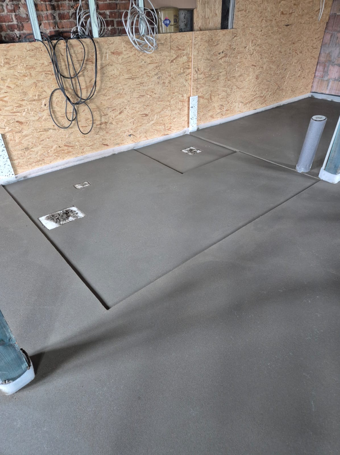 Świeżo wylana, gładka posadzka jastrychowa w surowym wnętrzu z widocznymi instalacjami elektrycznymi na ścianie z płyty OSB i fragmentem ceglanej ściany.