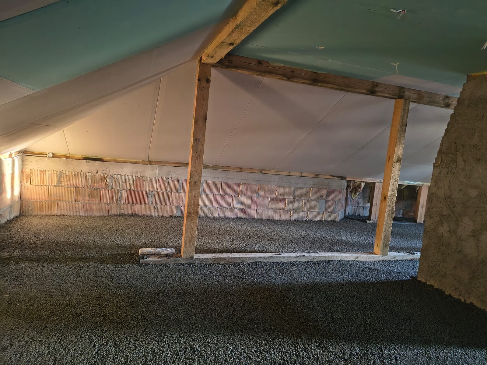 Szary jastrych na poddaszu z drewnianą konstrukcją, widoczne cegły na ścianie. Ujęcie z niskiego kąta, ukazujące strukturę wylewki i surowy stan wykończenia.