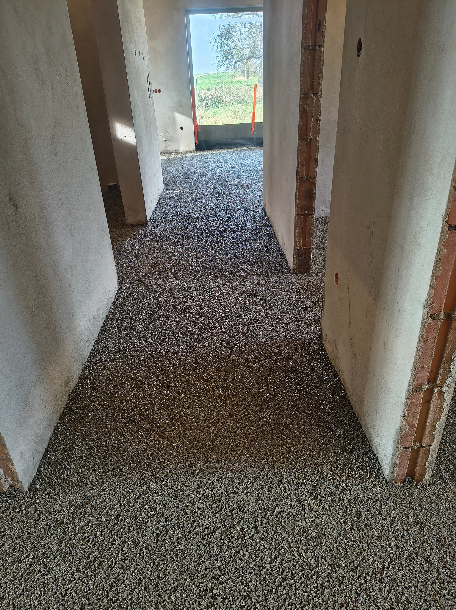 Wnętrze w trakcie prac wykończeniowych: świeżo wylana, szara posadzka jastrychowa z widocznymi fragmentami ścian i otworami drzwiowymi, w tle okno z widokiem na zieleń.