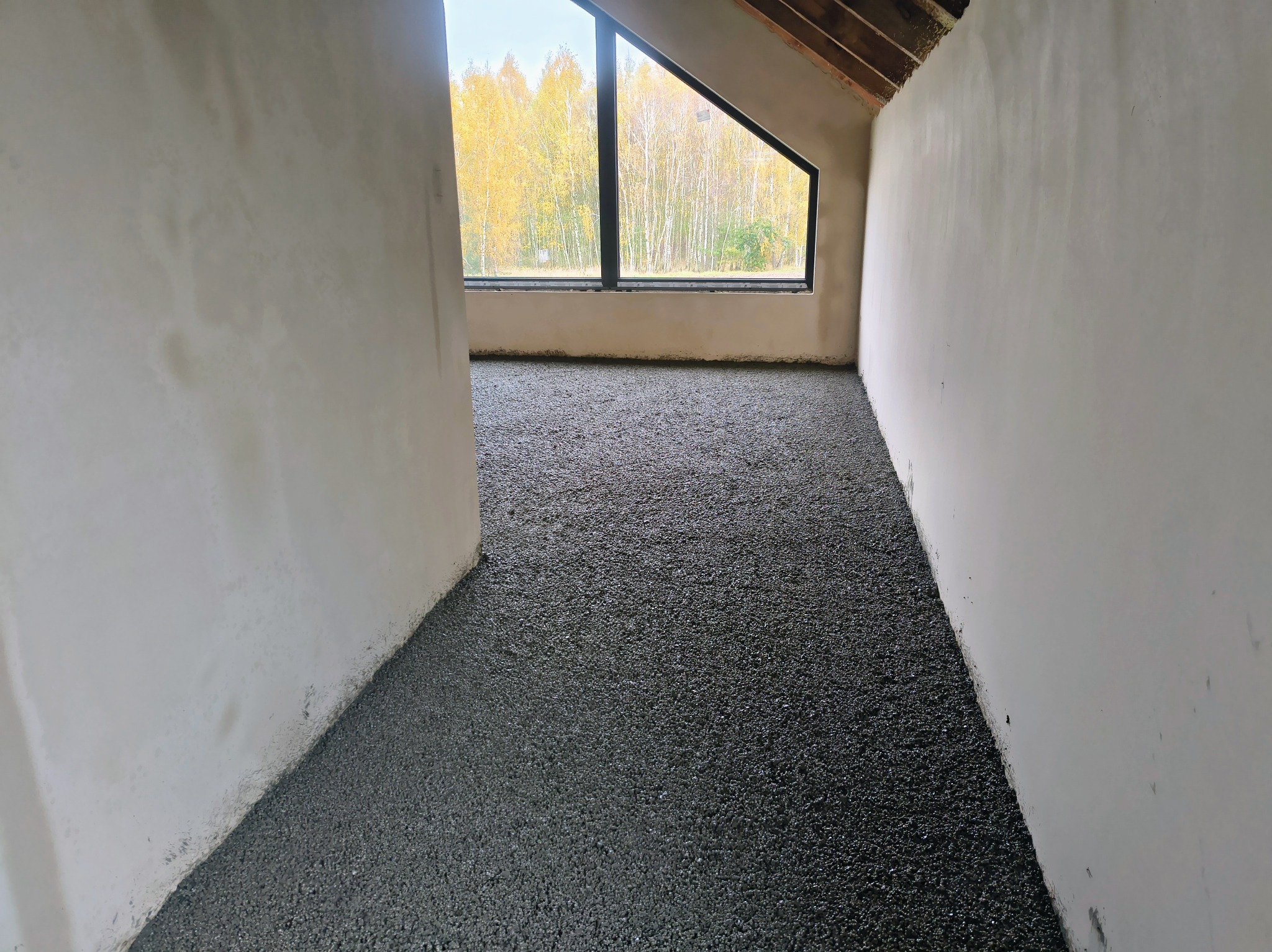 Świeżo wylana, ciemna posadzka w surowym wnętrzu z oknem na jesienny krajobraz. Widoczne ściany przygotowane do dalszych prac wykończeniowych.
