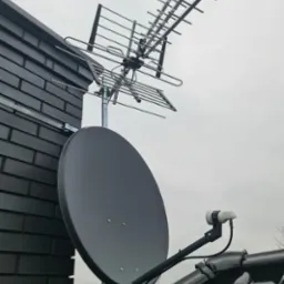 Antena satelitarna i antena naziemna zamontowane na dachu budynku z ciemnej cegły, widok z dołu na pochmurne niebo.