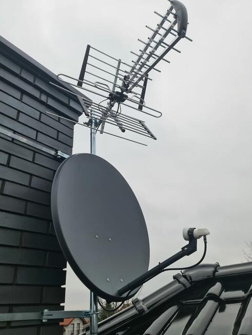 Antena satelitarna i antena naziemna zamontowane na dachu budynku z ciemnej cegły, widok z dołu na pochmurne niebo.