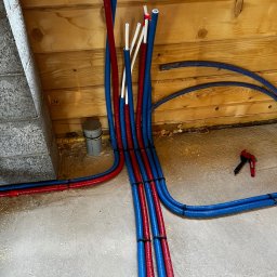 PRO INSTAL Piotr Ruciński - Instalacja wodna i grzewcza w podłodze, rury PEX w kolorach niebieskim i czerwonym, częściowo zamocowane do podłoża. Widoczne narzędzie do cięcia rur.