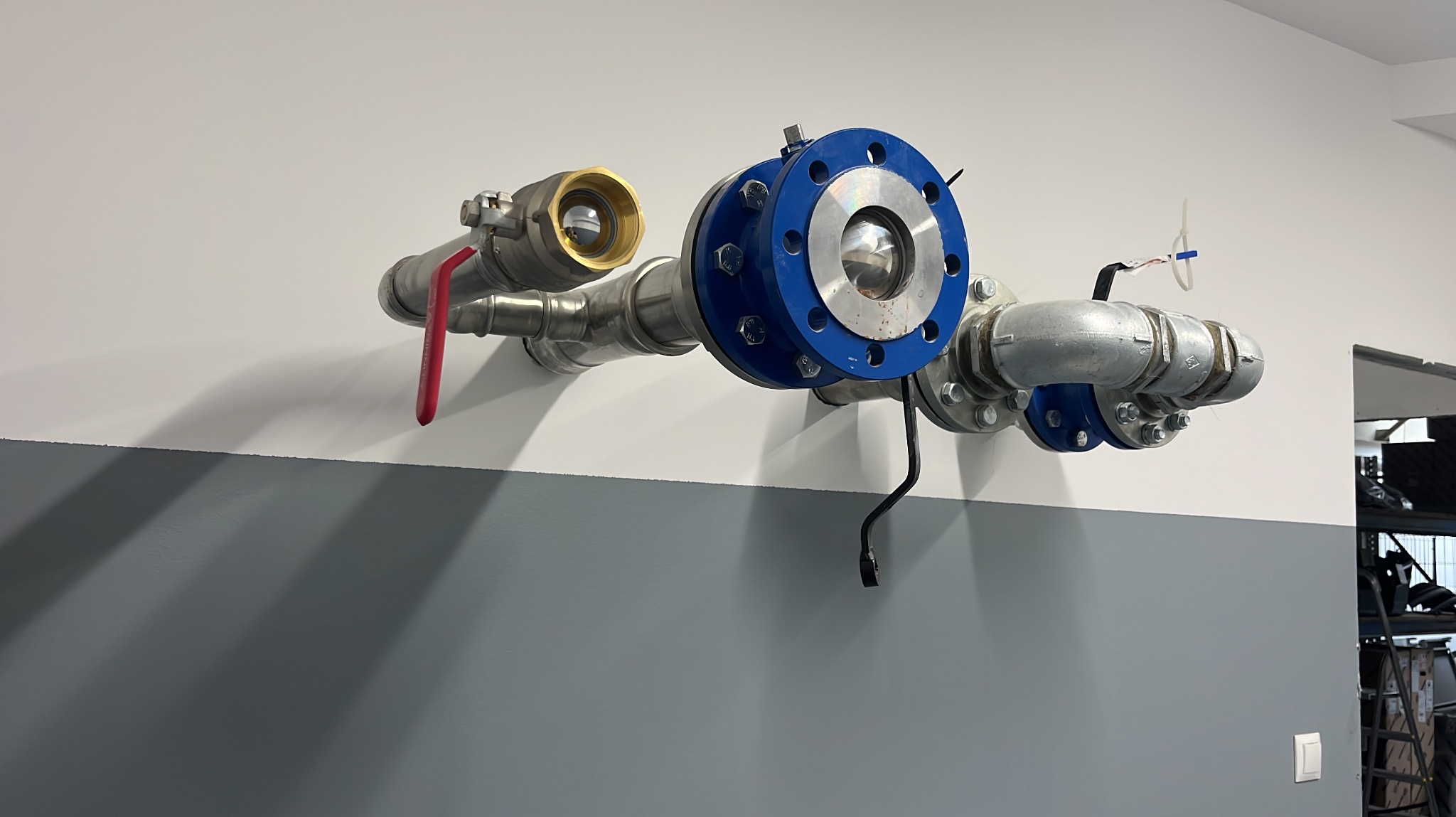 Metalowa instalacja rurowa z zaworem z czerwoną rączką i niebieskim kołnierzem, zamontowana na dwukolorowej ścianie. Widoczne śruby i złącza.
