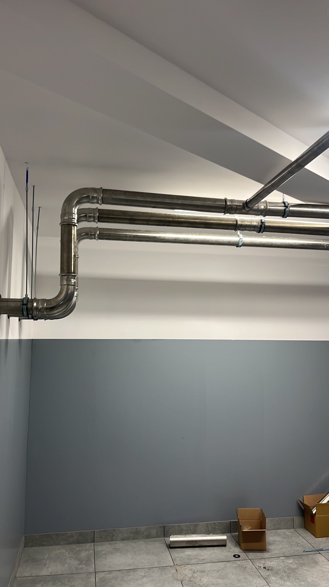 Równoległe, metalowe rury instalacyjne, zamocowane przy suficie w pomieszczeniu z szarą ścianą. Widoczne złączenia i mocowania rur.