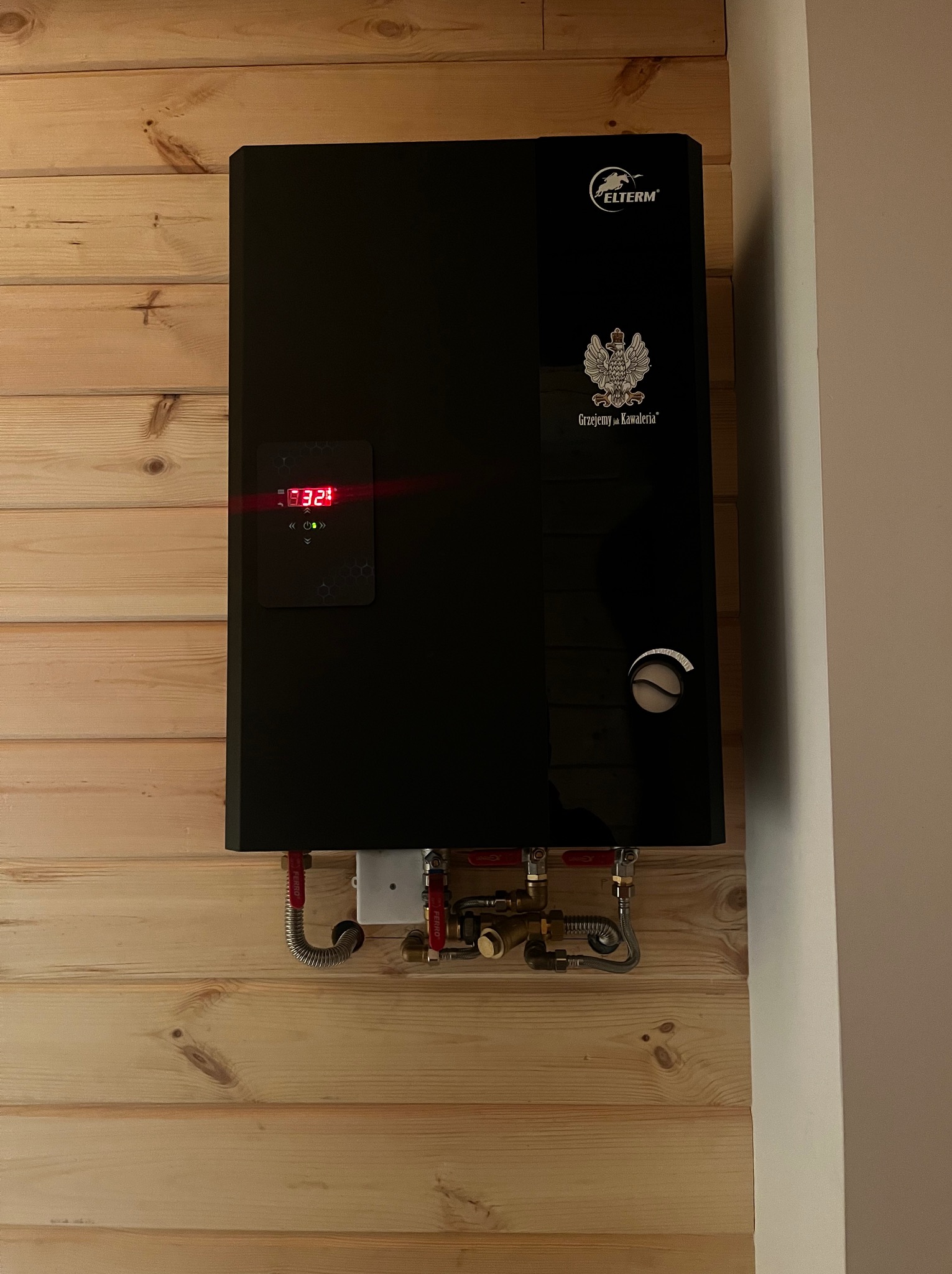 Czarny piec gazowy Elterm z wyświetlaczem i armaturą, zamontowany na drewnianej ścianie. Widoczne przyłącza i logo producenta.