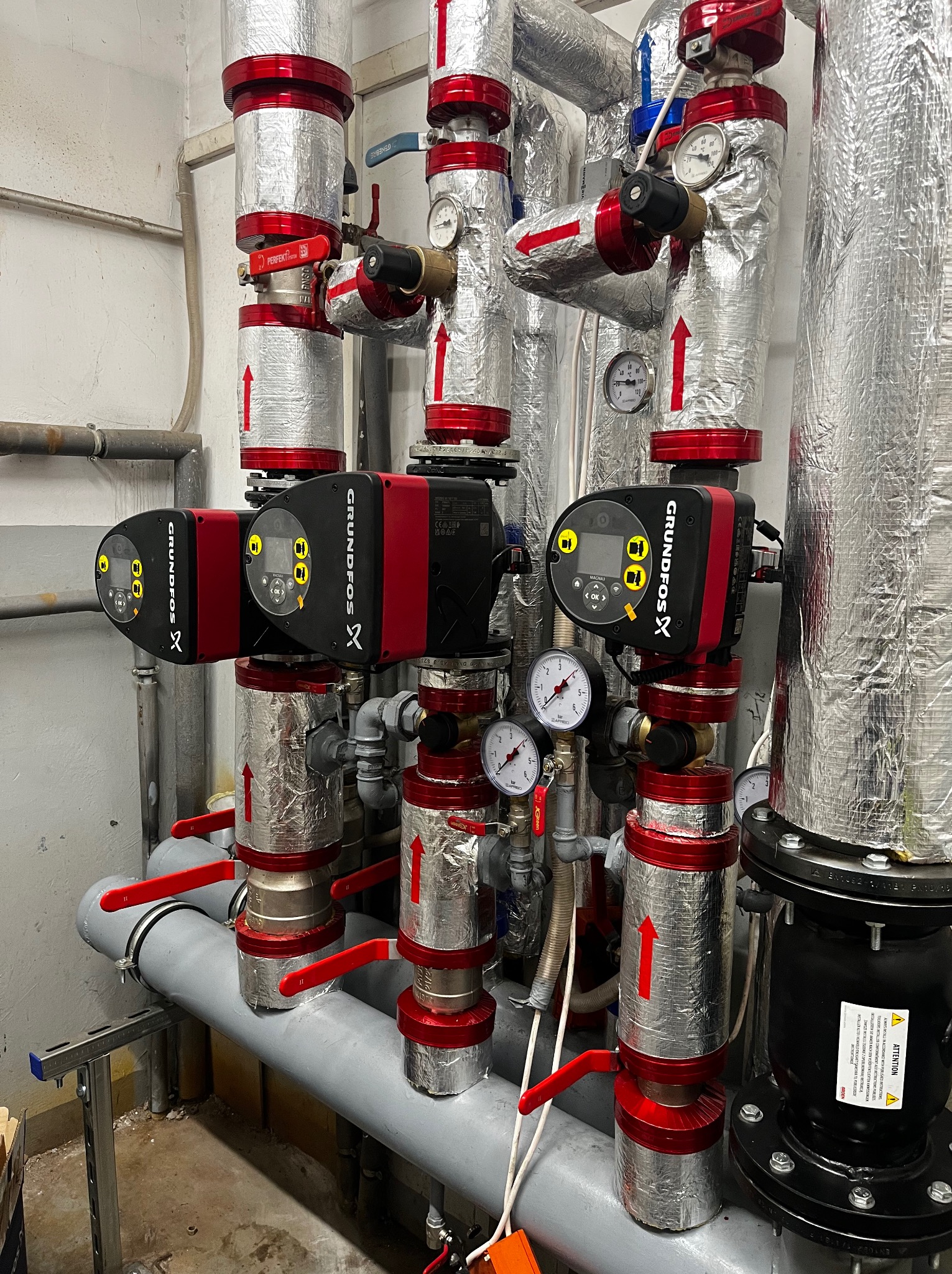 Złożona instalacja grzewcza z pompami obiegowymi Grundfos, rurami w izolacji termicznej z czerwonymi elementami i zaworami. Widoczne manometry i zawory.