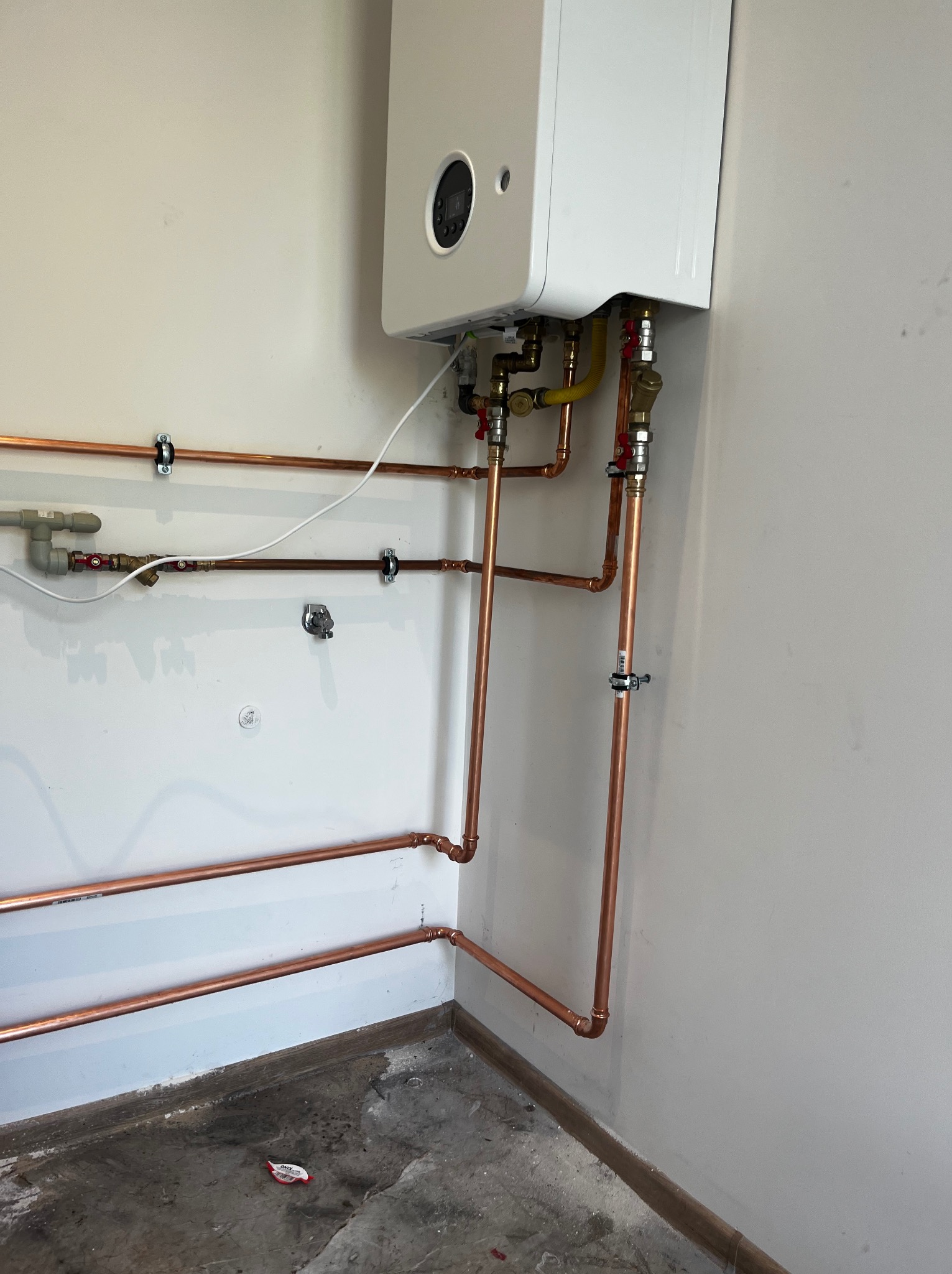 Biały piec gazowy na ścianie z miedzianymi rurami instalacji, widoczne zawory i mocowania. Surowy beton na podłodze. Ujęcie z dołu, instalacja po montażu.