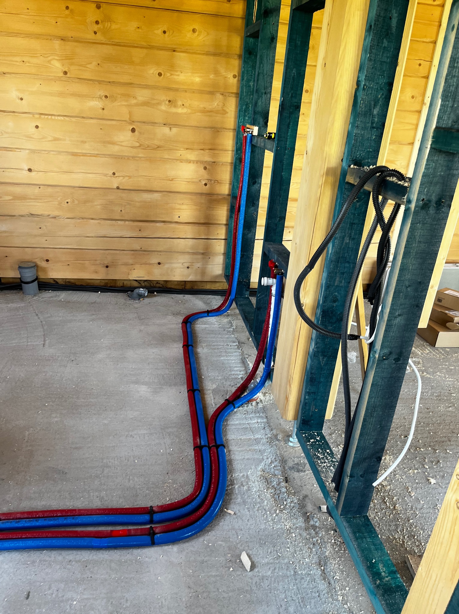 Instalacja wodna w budynku szkieletowym: rury PEX (czerwone i niebieskie) prowadzone wzdłuż drewnianej ściany, przymocowane czarnymi opaskami. Widoczne elementy konstrukcji i podłoże.