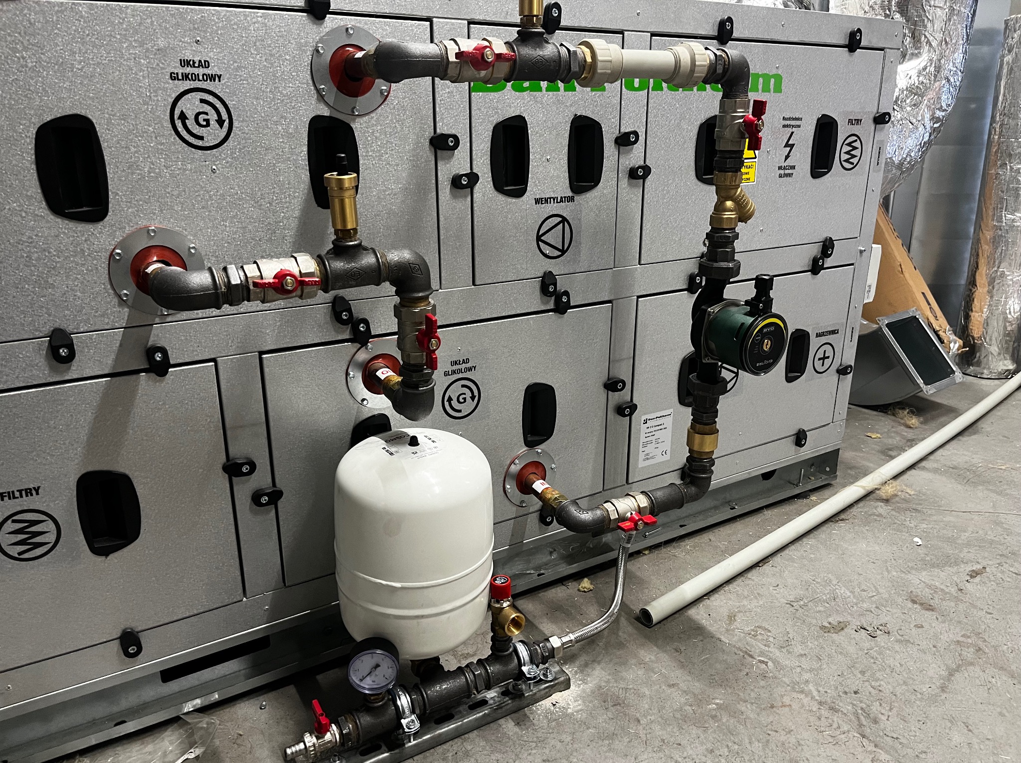 Złożona instalacja wentylacyjna z widocznymi elementami hydraulicznymi, w tym pompą obiegową i zbiornikiem wyrównawczym, na tle szarej podłogi.