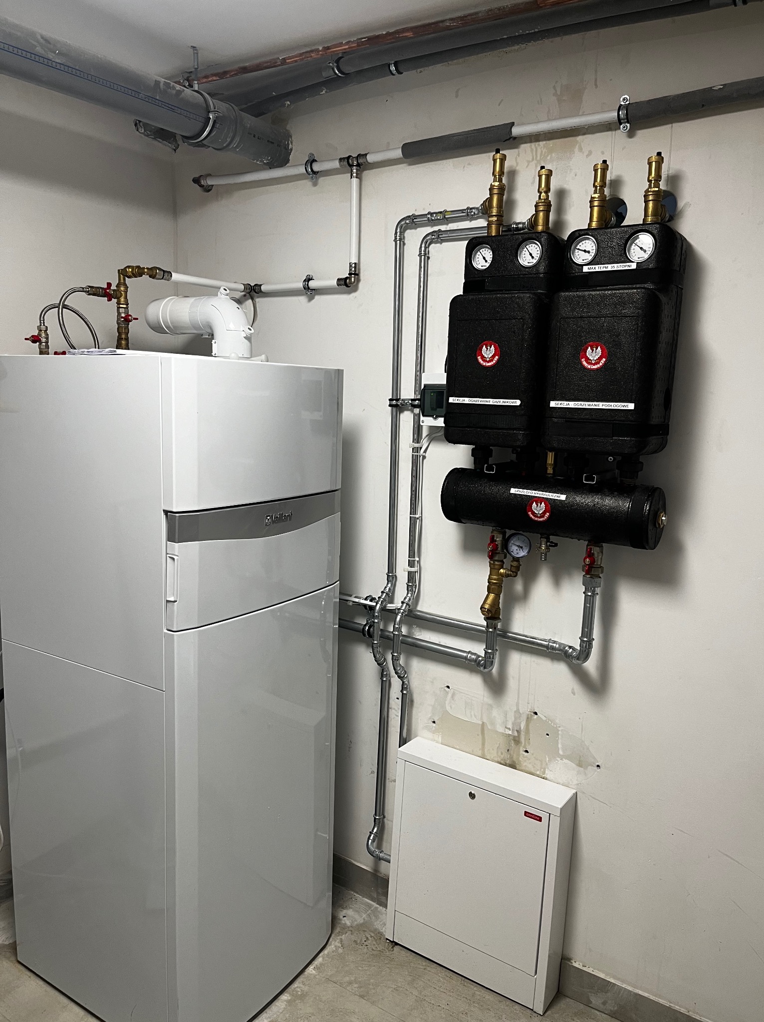 Biały piec grzewczy Vaillant z podłączonymi rurami i czarnymi modułami sekcji grzejnikowej i podłogowej. Widoczna skrzynka instalacyjna na ścianie.