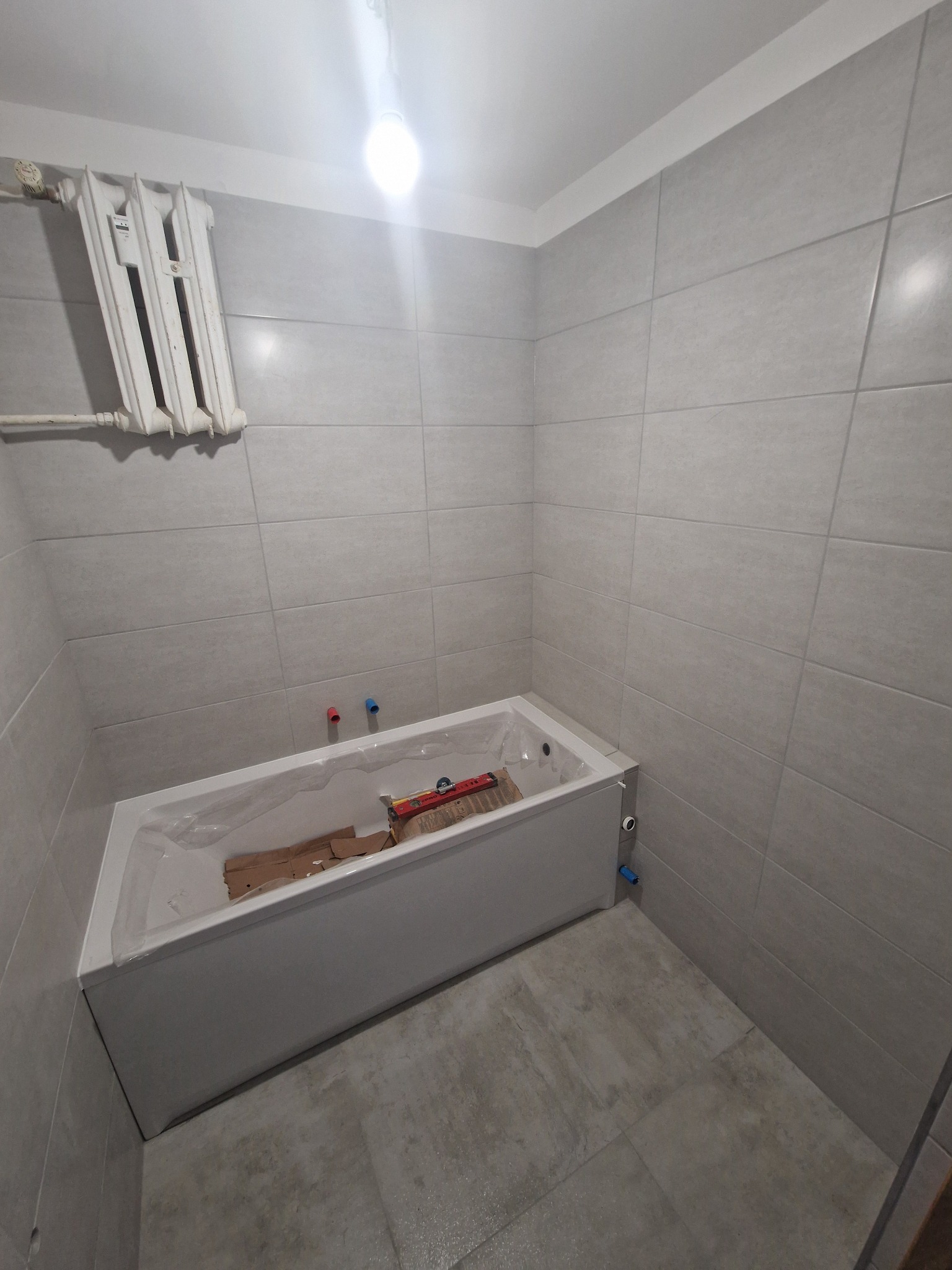 Nowa, biała wanna w trakcie montażu w wykafelkowanej, szarej łazience; widoczne narzędzia i kartony zabezpieczające, grzejnik na ścianie.