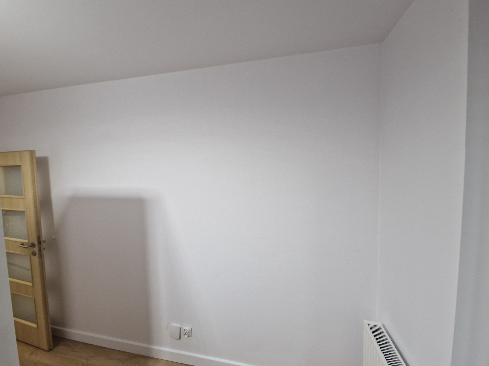 Pomalowane na biało wnętrze pokoju z drewnianymi drzwiami z przeszkleniami po lewej stronie, widoczne gniazdka elektryczne przy podłodze i grzejnik pod ścianą.