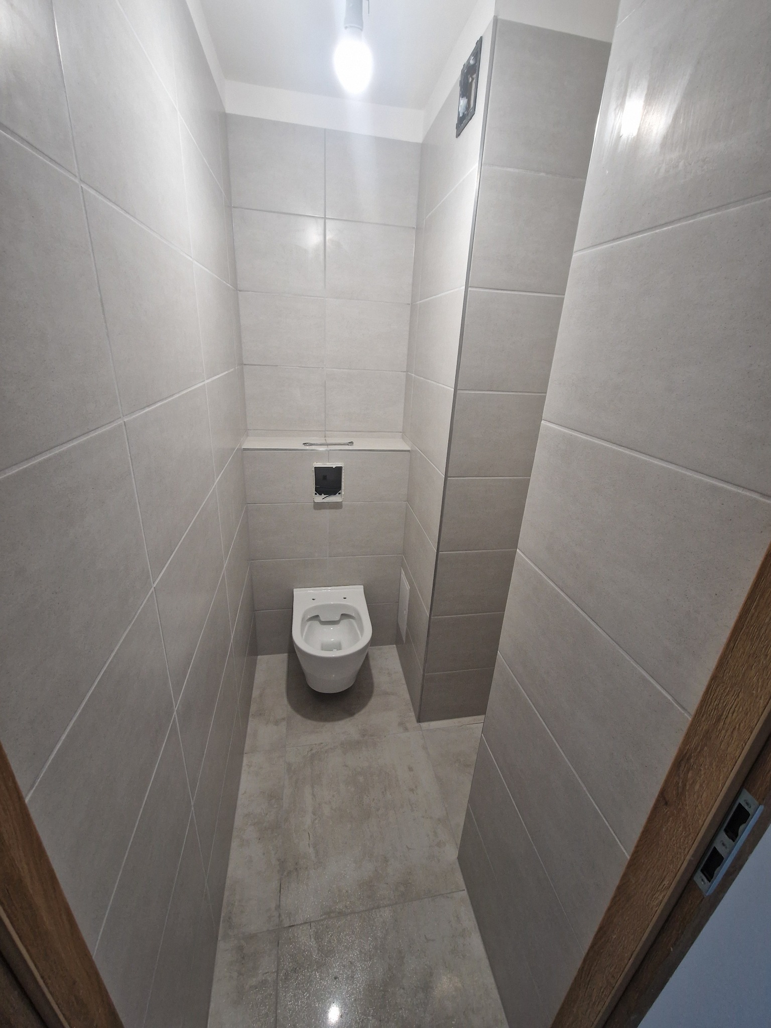 Mała łazienka z szarymi, matowymi płytkami na ścianach i podłodze, wisząca miska WC i widoczna instalacja podtynkowa, oświetlenie górne.