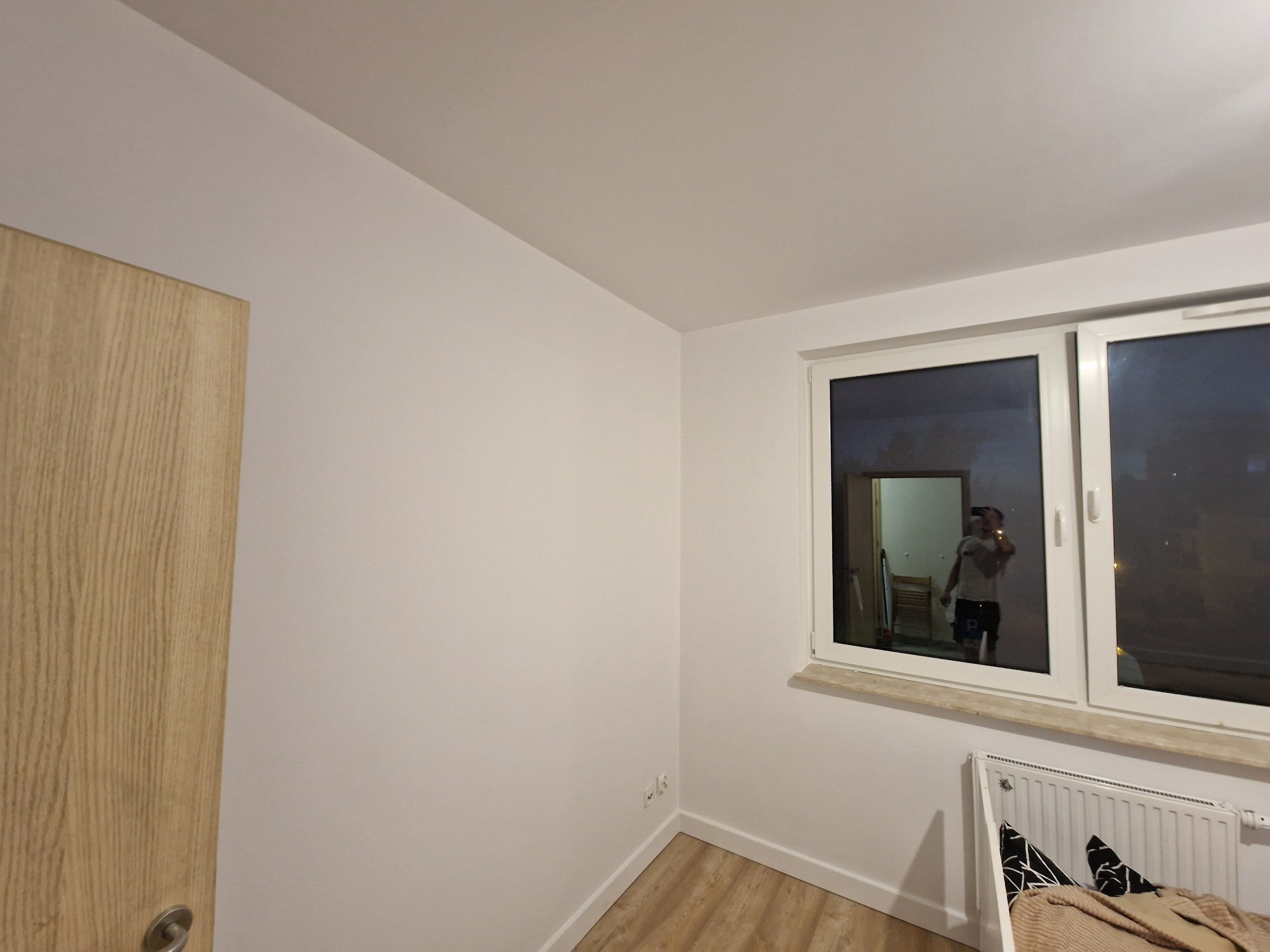 Pomalowane na biało wnętrze pokoju z drewnianą podłogą, grzejnikiem pod oknem i odbiciem osoby w szybie okiennej.