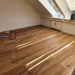 S line - Poddasze z oknami dachowymi w trakcie układania paneli podłogowych. Widoczne panele, karton, narzędzia i grzejniki. Jasne światło dzienne.