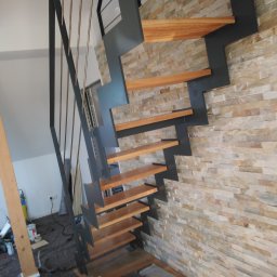 S line - Nowoczesne schody z drewnianymi stopniami i metalową konstrukcją, zamontowane przy ścianie wyłożonej dekoracyjnym kamieniem. Widoczne materiały budowlane w tle.
