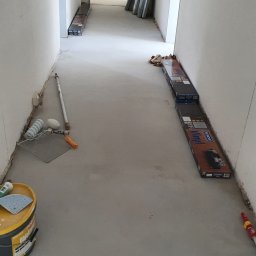 S line - Przygotowanie do prac w korytarzu w Głogowie: puszka z farbą, wałki malarskie i panele podłogowe ułożone wzdłuż ścian. Widoczny surowy beton i drzwi w tle.