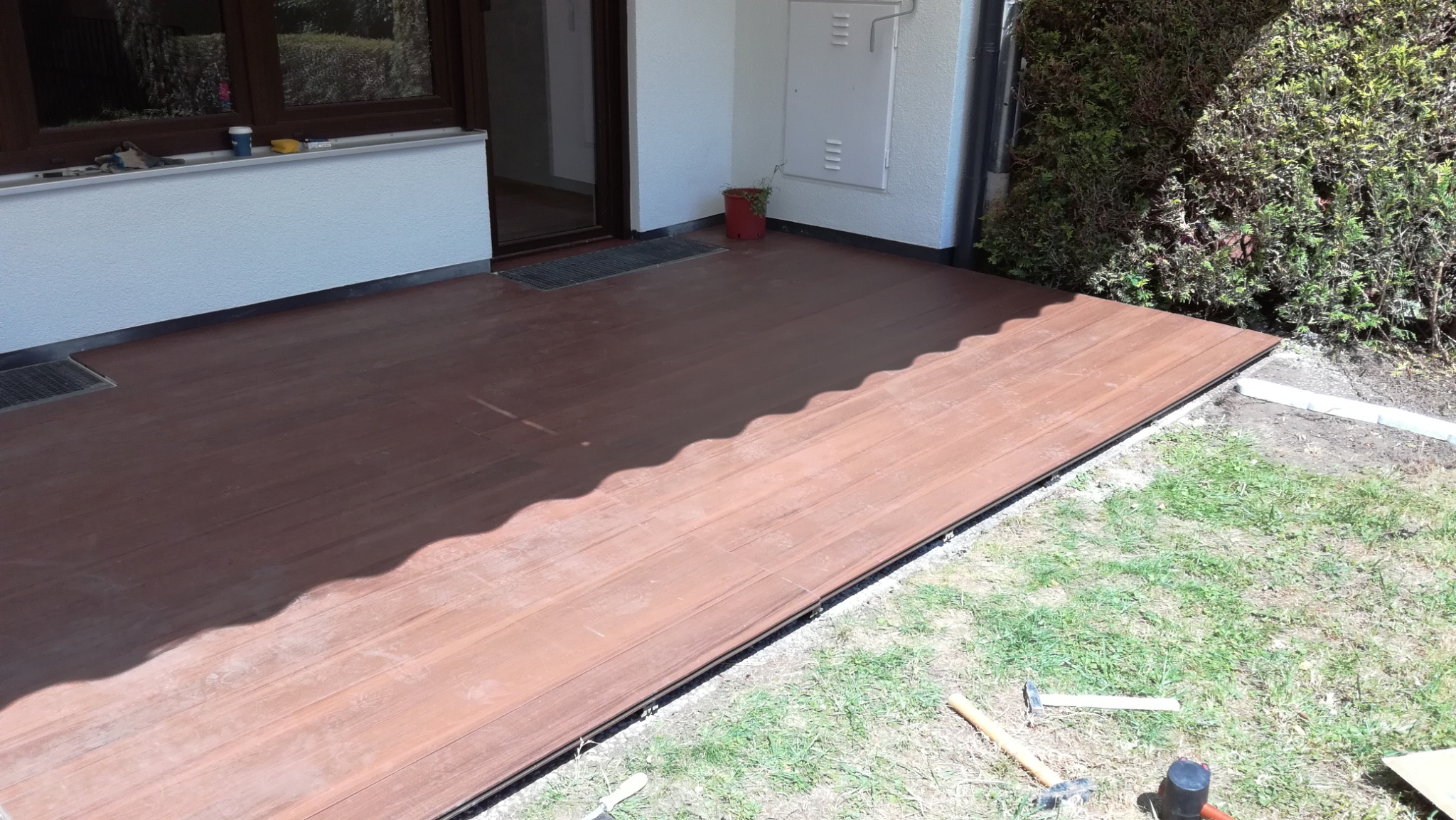 Brązowy taras z kompozytowych desek, częściowo zacieniony, z widocznymi narzędziami i fragmentem zieleni. Detal wykończenia przy budynku.