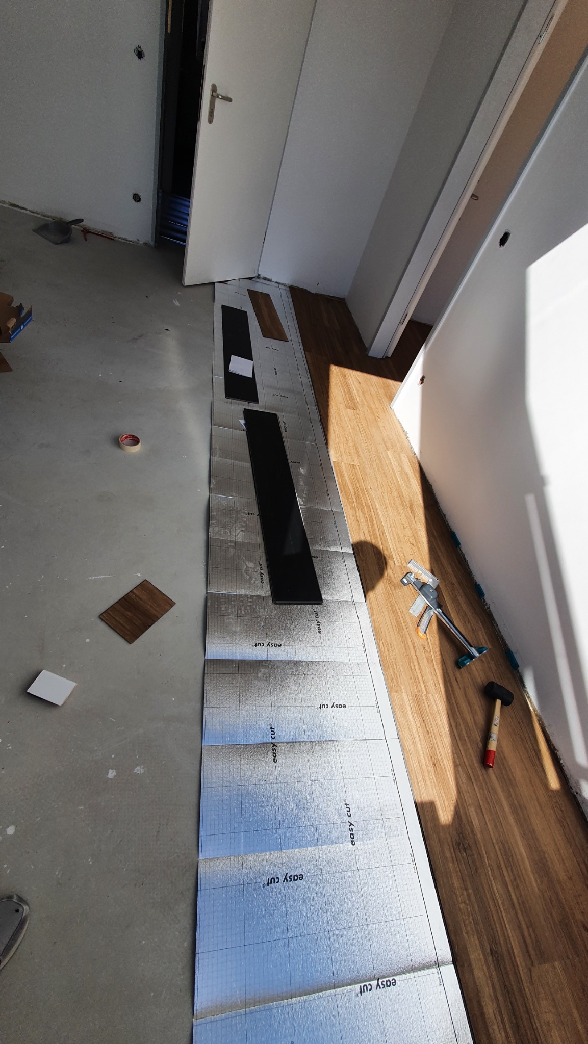 Fragment podłogi w trakcie układania paneli. Widoczna folia izolacyjna, narzędzia i częściowo ułożone panele w jasnym pomieszczeniu.