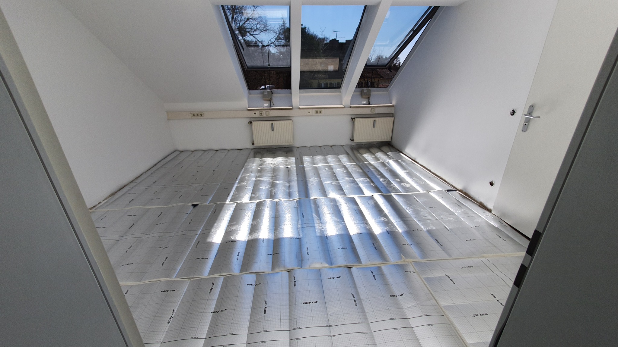 Przygotowanie podłoża pod panele w pokoju na poddaszu. Widoczne okna dachowe, grzejniki i rozłożona folia izolacyjna z nadrukiem 'easy cut'.