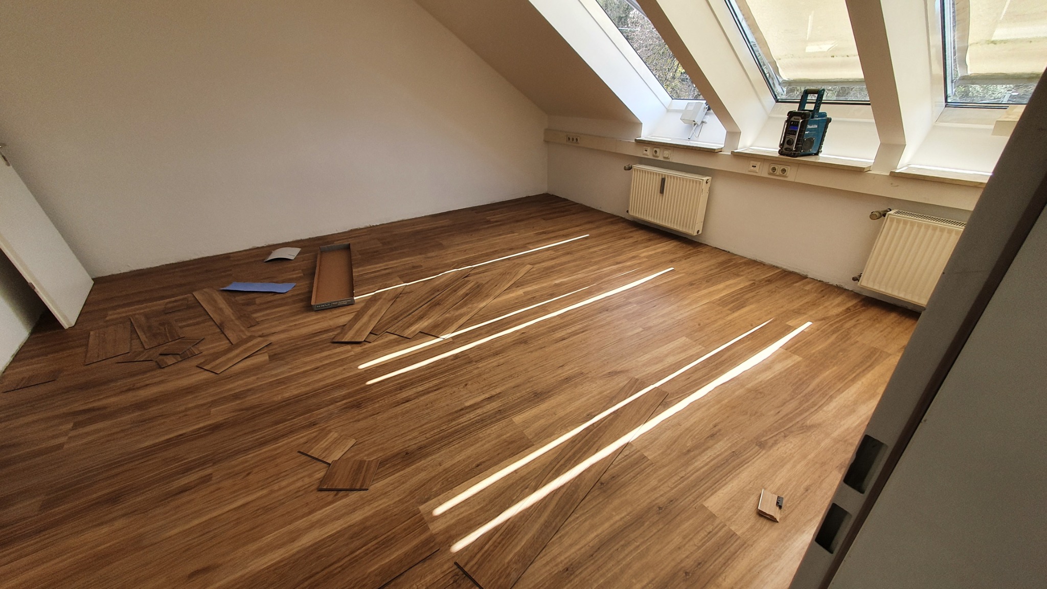 Poddasze z oknami dachowymi w trakcie układania paneli podłogowych. Widoczne panele, karton, narzędzia i grzejniki. Jasne światło dzienne.