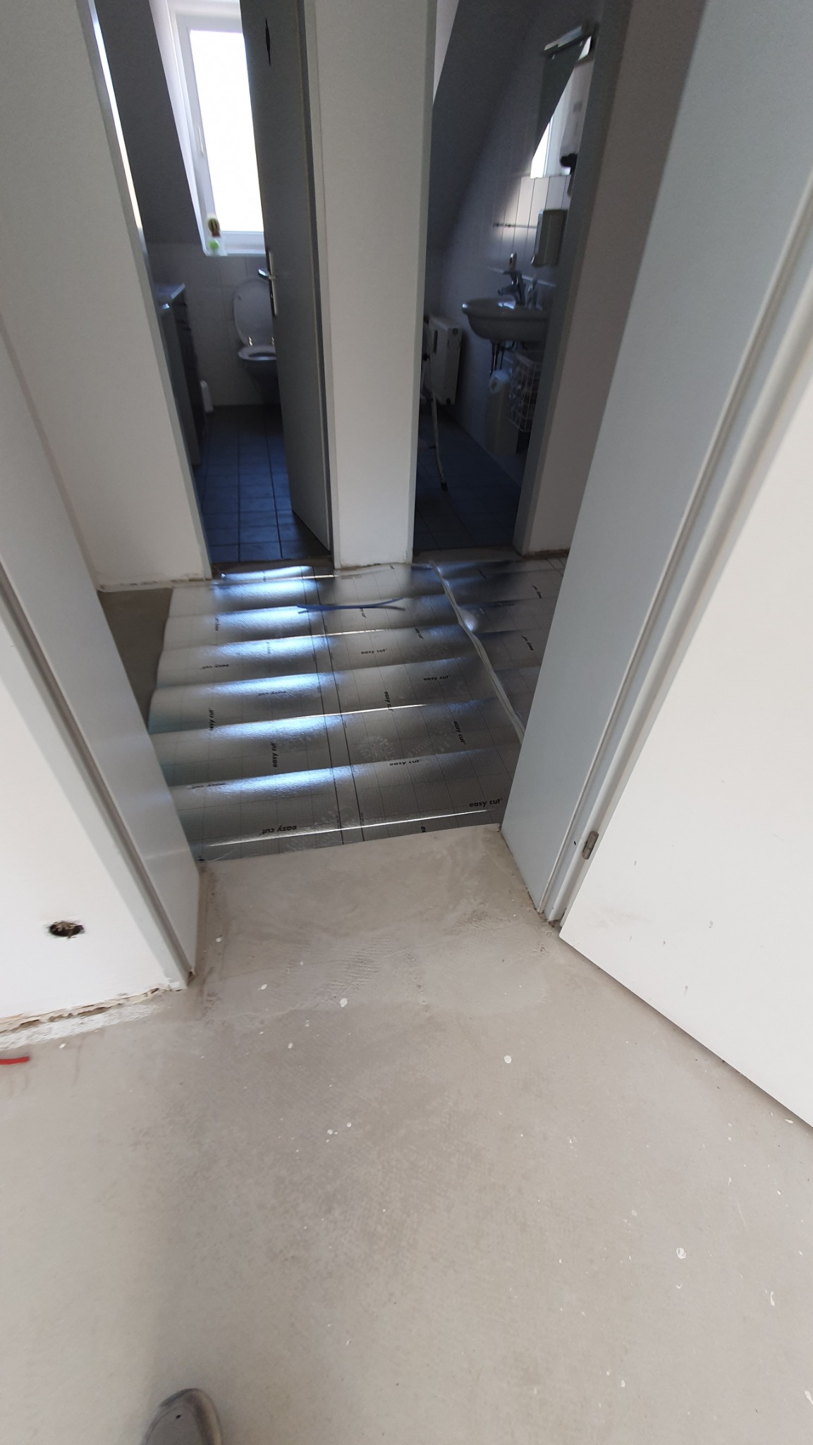 Przygotowanie podłoża pod panele w łazience na poddaszu: rozłożona folia izolacyjna na wylewce, widok przez otwarte białe drzwi do toalety i łazienki.