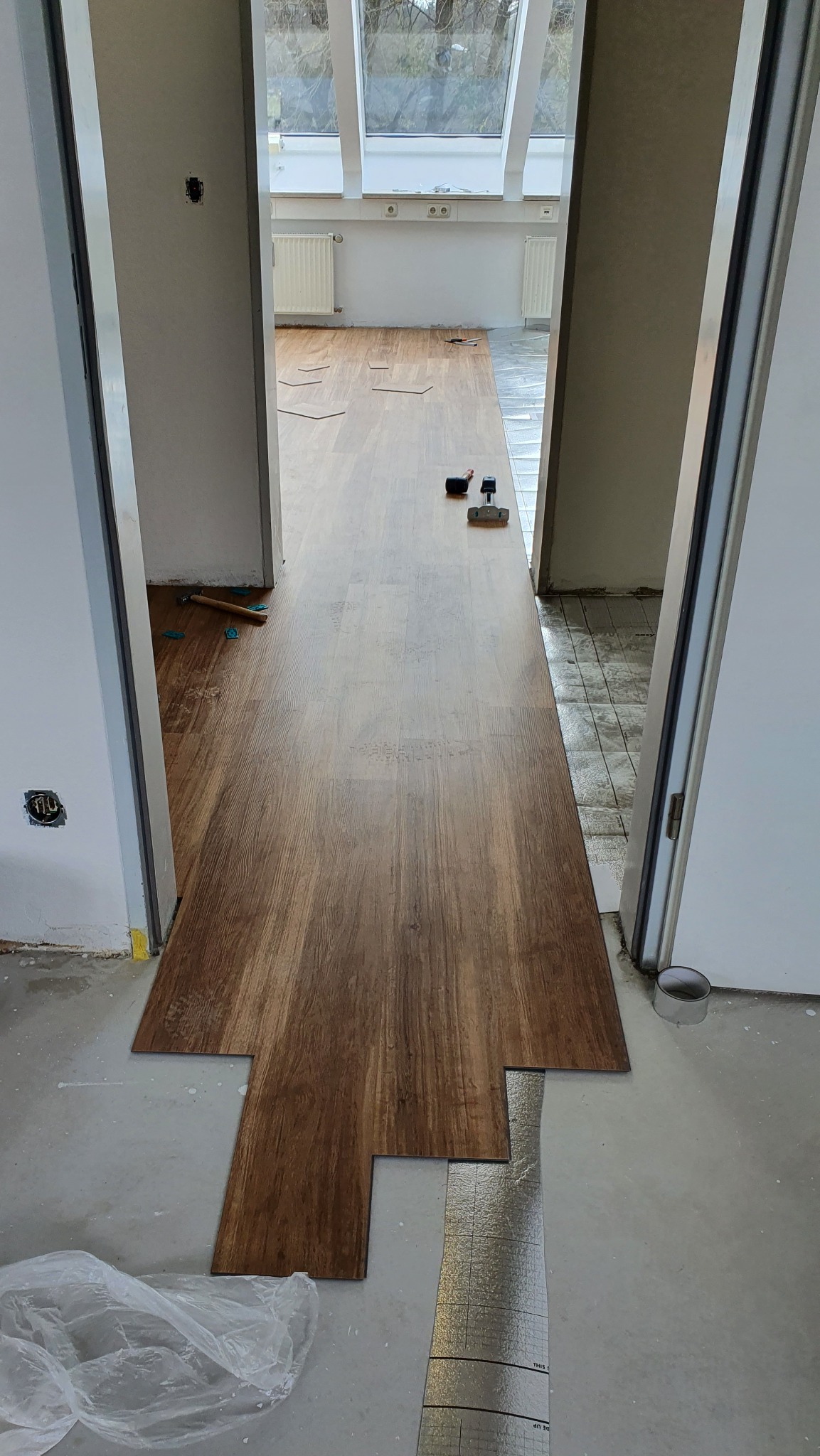 Układanie paneli podłogowych w jasnym pomieszczeniu z oknami dachowymi. Widoczne narzędzia i podkład. Częściowo ułożona podłoga z paneli imitujących drewno.